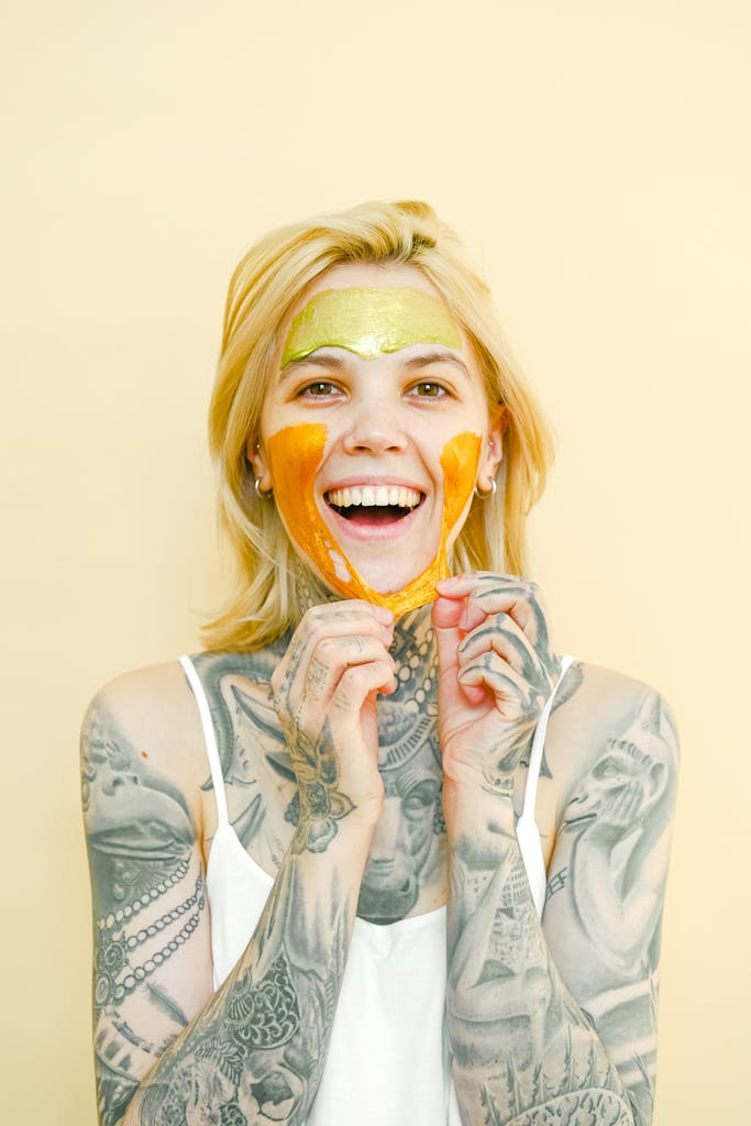 Young cheerful female with tattooed body and blond hair removing peel off golden mask from face looking at camera