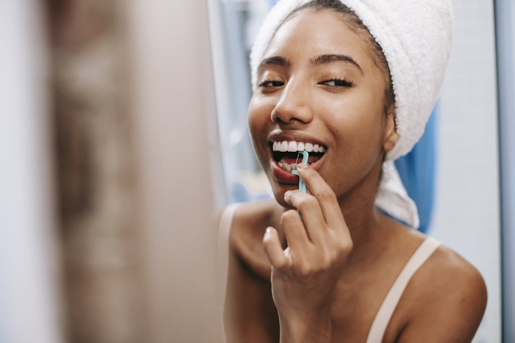 Smiling African American female with white towel on head cleaning teeth with dental flosser in bathroom