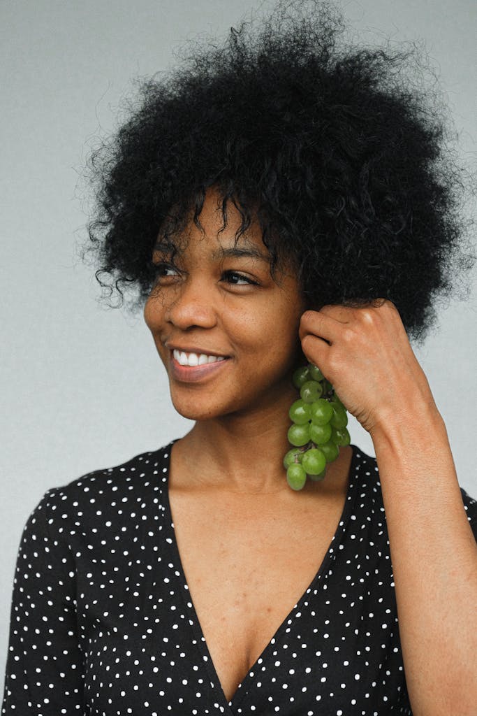 Charming black female model in polka dot dress holding bunch of grapes like earring looking away with smile